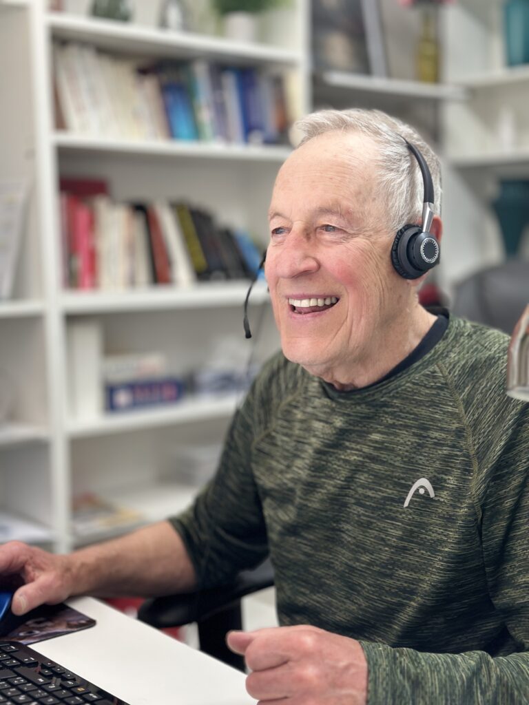 Smiling older man wearing a headset while using a computer at a desk in a home office