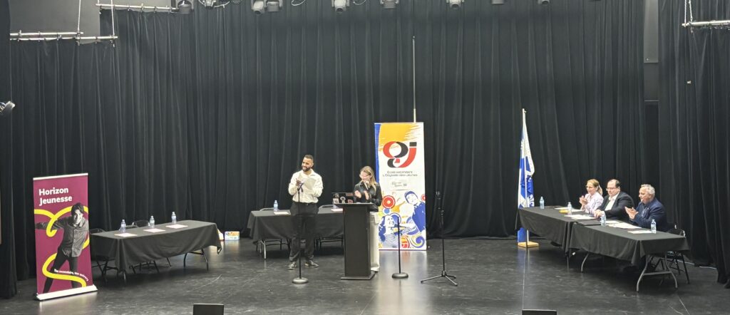 Man and woman at a black podium presenting on a stage, with a banner and a panel of five seated at long tables on the right side.