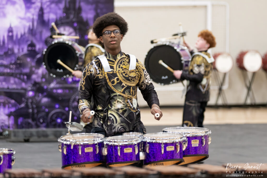 Young drummer with afro and glasses playing a row of purple drums in a black-gold uniform, with other drummers in the background.