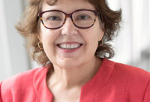Smiling woman with short brown hair and red glasses, wearing a coral blazer and white top, portrait indoors.