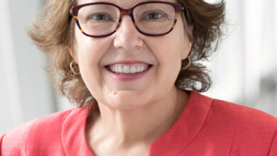 Smiling woman with short brown hair and red glasses, wearing a coral blazer and white top, portrait indoors.