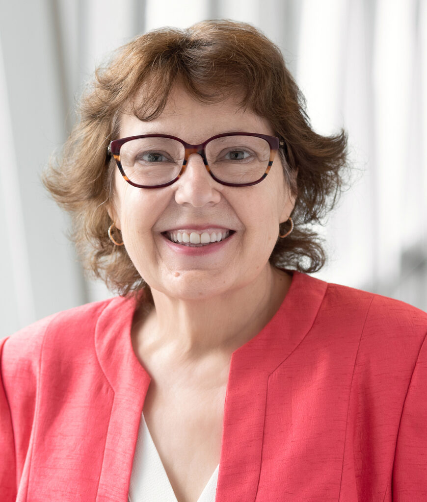 Smiling woman with short brown hair and red glasses, wearing a coral blazer and white top, portrait indoors.