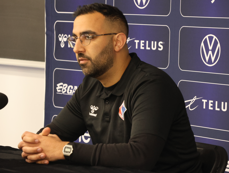 Man with glasses and a trimmed beard sits at a table during a press conference, hands folded in front of him.