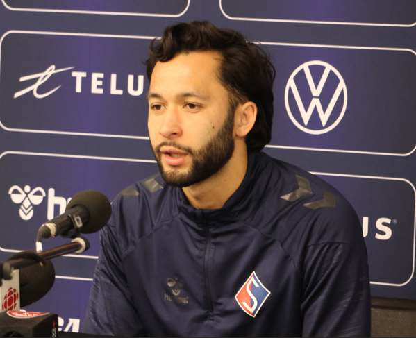 Man with dark hair and beard speaks at a press conference, wearing a navy jacket with a crest; microphone in front and sponsor logos in the background.