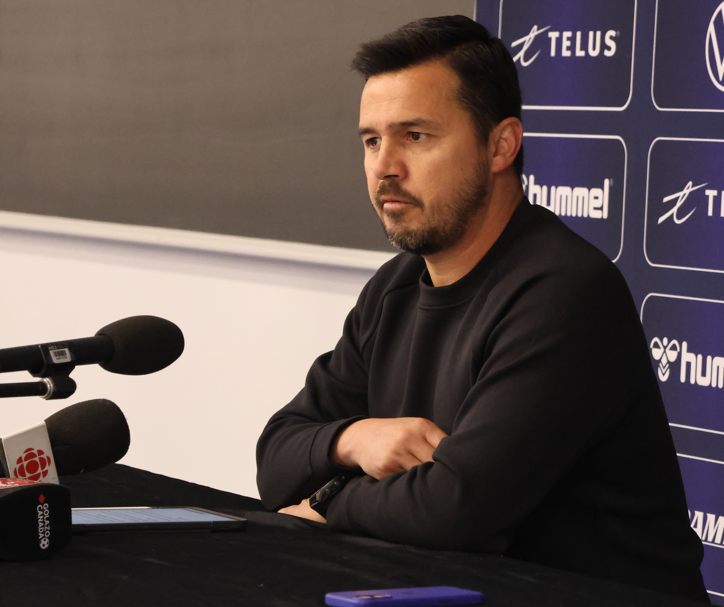 Man in a black sweater sits at a desk during a press conference, arms crossed, microphones in front, blue sponsor backdrop behind.