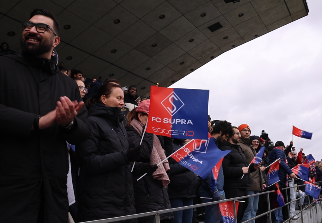 Crowd of football fans in winter coats holding red and blue FC Supra flags at a stadium stands.