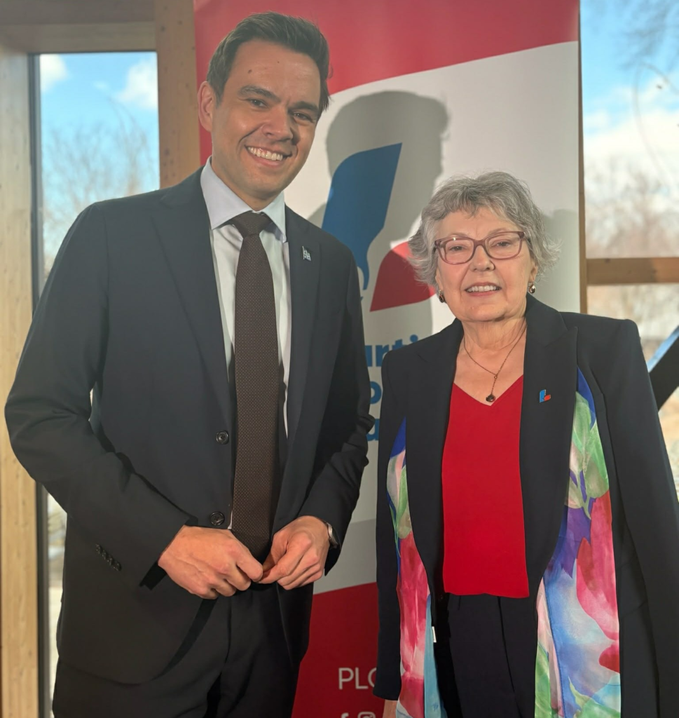 Man in a suit posing with an older woman in a red blouse and colorful scarf at an event backdrop.