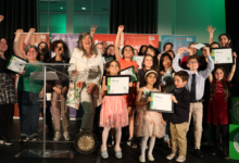 Group of children and adults on a stage, holding framed certificates and cheering during an awards ceremony.