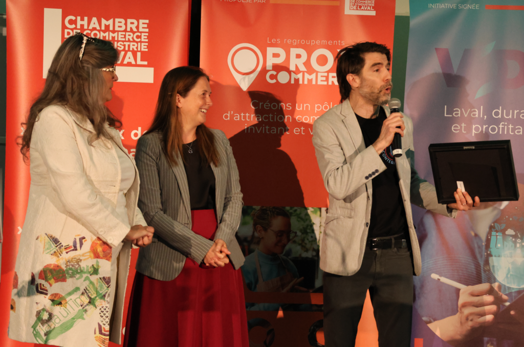 Three adults on a stage: a man with a microphone holds a framed award, flanked by two smiling women, with red and blue banners behind them.