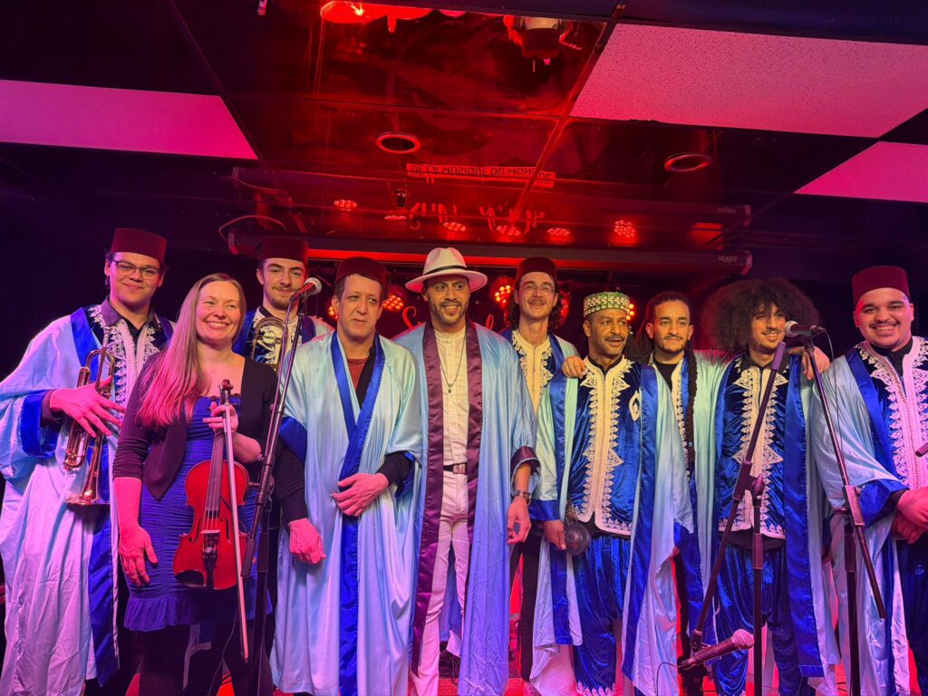 Group of performers in blue robes and fez hats on a stage, posing with instruments under red lighting.