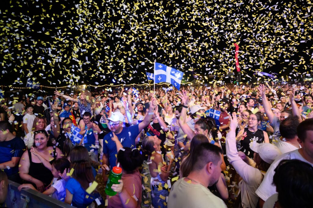 Large nighttime crowd celebrating with yellow and white confetti falling from above; many people raise hands and wave Quebec flags.