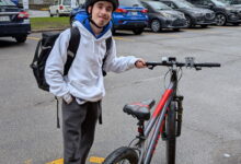 Young person wearing a helmet and backpack standing next to a red-and-gray mountain bike in a parking lot with yellow lines and leafless trees in the background