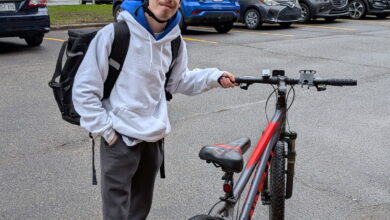 Young person wearing a helmet and backpack standing next to a red-and-gray mountain bike in a parking lot with yellow lines and leafless trees in the background