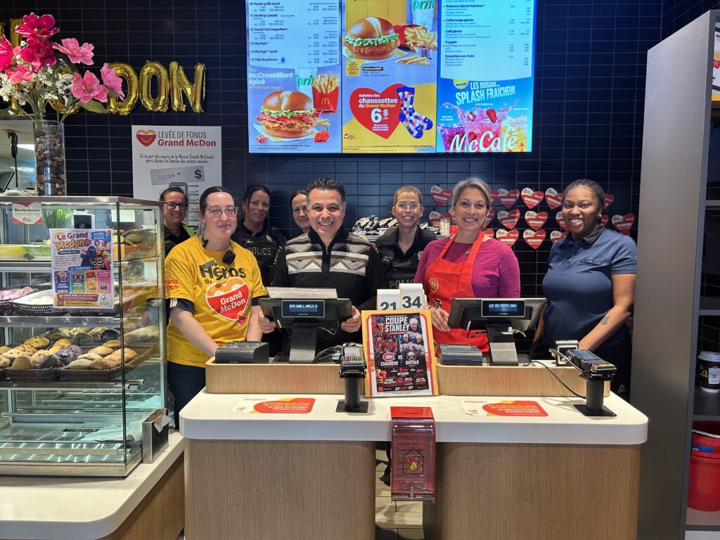 Group of McDonald's employees behind the counter, smiling at the camera with cash registers and digital menu boards above them.