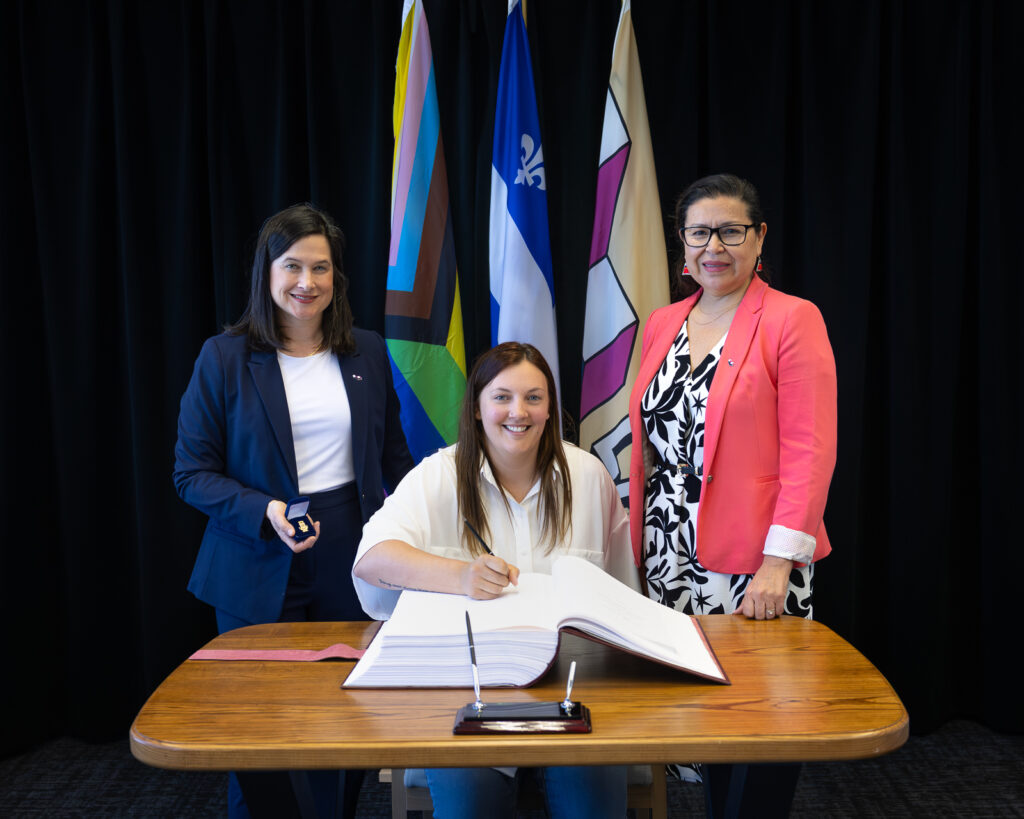 SignaturelivredorLaval Média Laval Woman seated at a wooden table signs a document, flanked by two colleagues in blazers with colorful flags in the background.