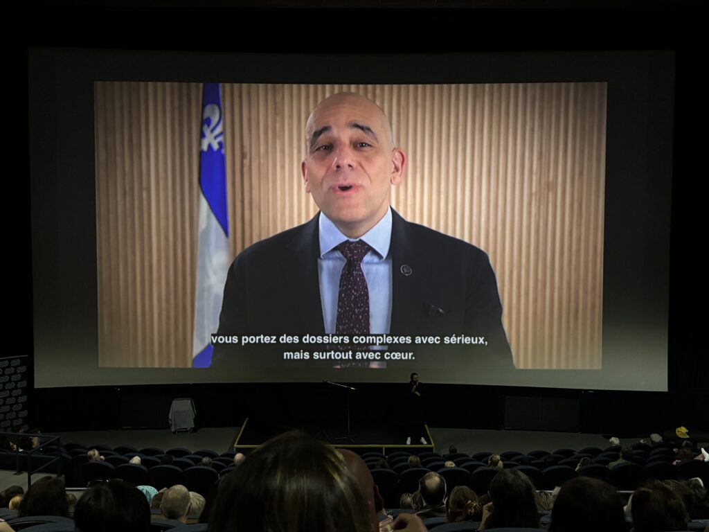 Skeete Média Laval Man in a suit speaking on a large screen in a theater, with a striped wooden backdrop and a blue flag behind him, subtitles visible at the bottom in French.