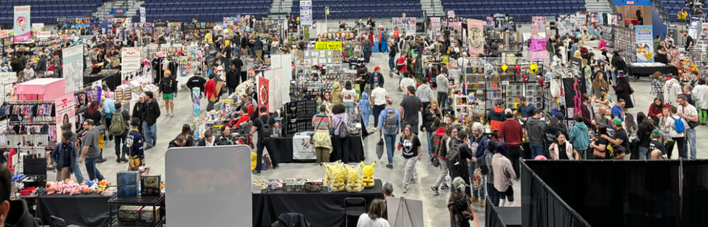 Large indoor convention hall filled with numerous vendor booths and shoppers browsing merchandise on displays and tables in a wide aisles layout.