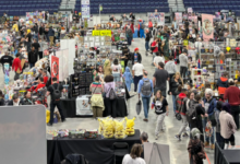 Large indoor convention hall filled with numerous vendor booths and shoppers browsing merchandise on displays and tables in a wide aisles layout.