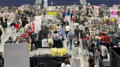 Large indoor convention hall filled with numerous vendor booths and shoppers browsing merchandise on displays and tables in a wide aisles layout.