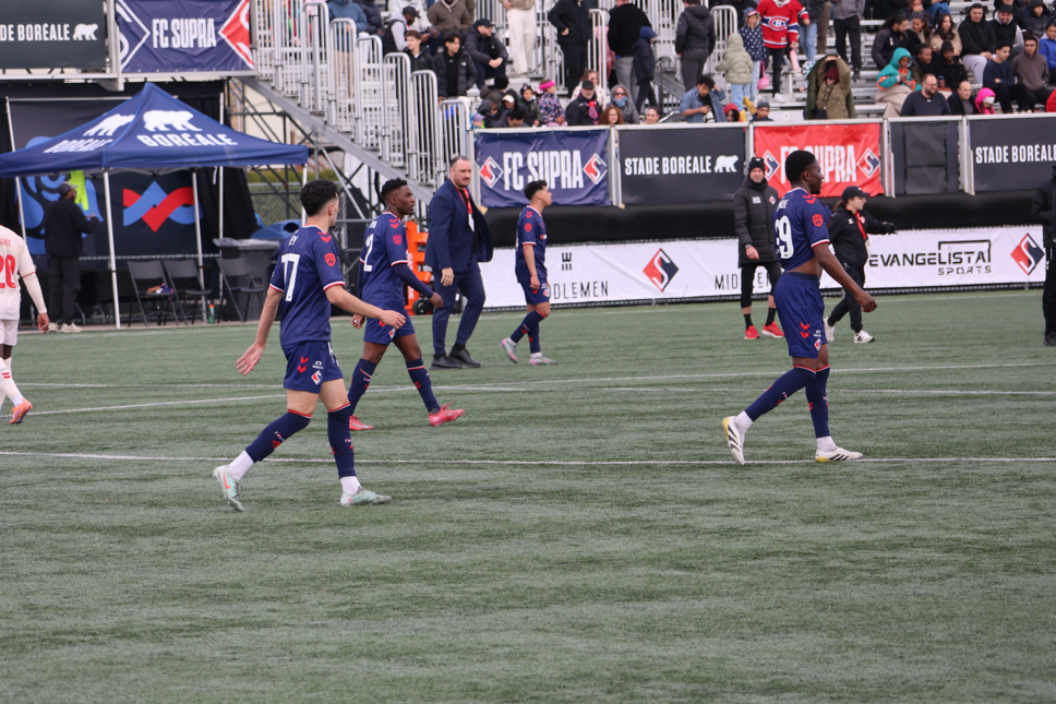 Soccer players in blue kits walk across a green field with spectators in the stands behind them and banners along the sideline.