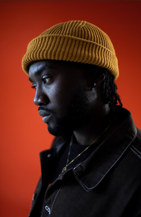 Male portrait in profile wearing a mustard knit beanie and dark denim jacket against an orange background.