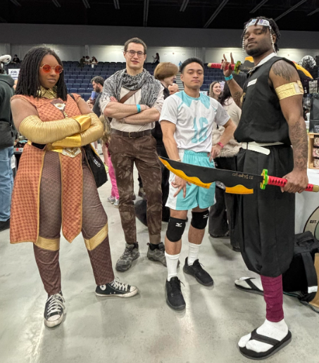 Group of cosplayers posing at a convention; a woman in orange armor and red sunglasses stands left, three men in varied costumes center, and a tall man in black with a yellow sword prop on the right.