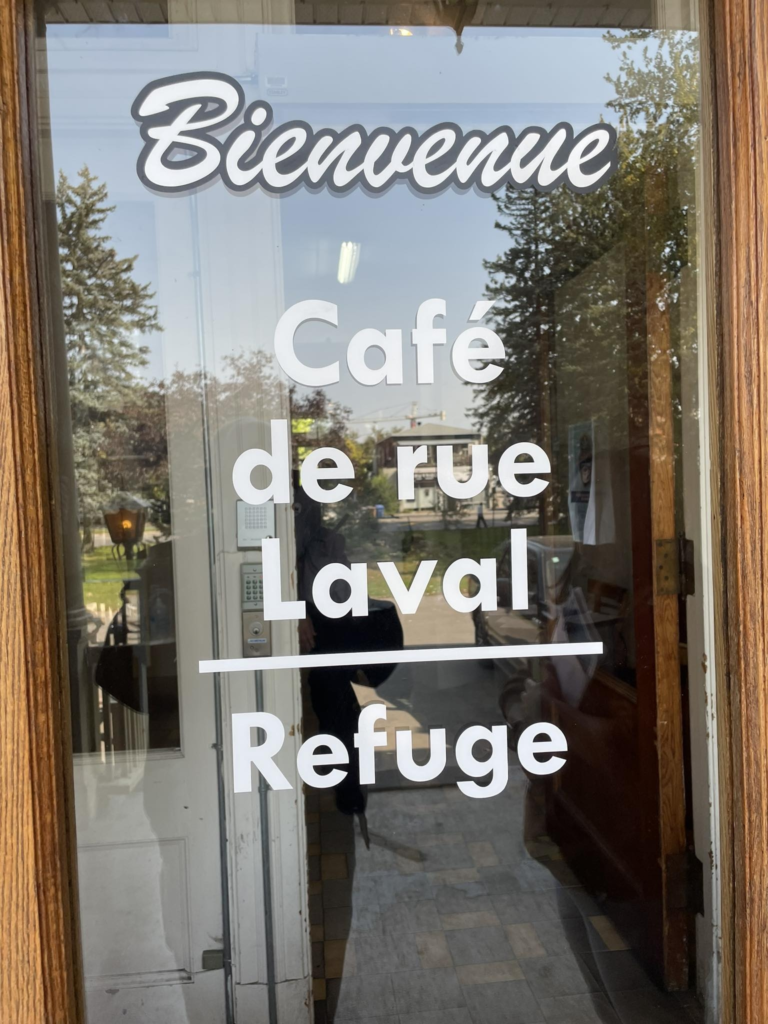 image Média Laval Glass door with white sign reading 'Bienvenue Café de rue Laval Refuge' and a wooden frame outside.