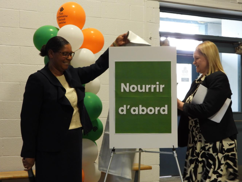 Two women in business attire attach a green-framed sign reading 'Nourrir d'abord' beside a balloon column.
