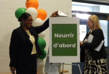 Two women in business attire attach a green-framed sign reading 'Nourrir d'abord' beside a balloon column.