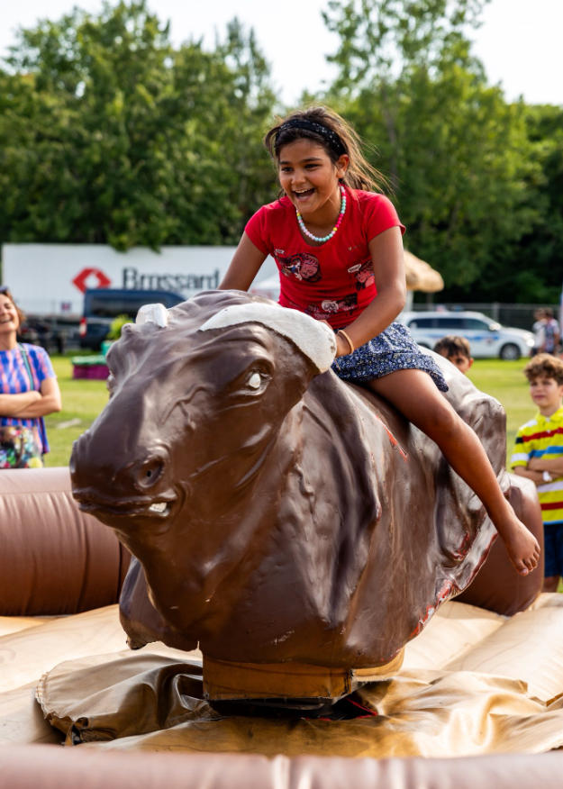 Girl in a red shirt rides a mechanical bull at an outdoor fair, smiling with spectators in the background