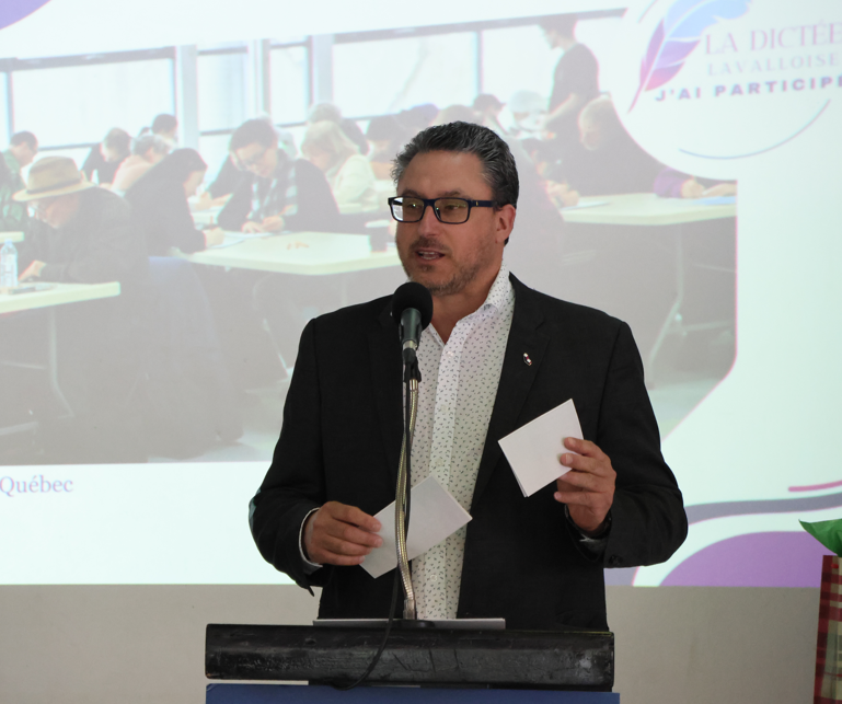 Man in a dark blazer and glasses speaks at a podium with a microphone, holding papers during a presentation.