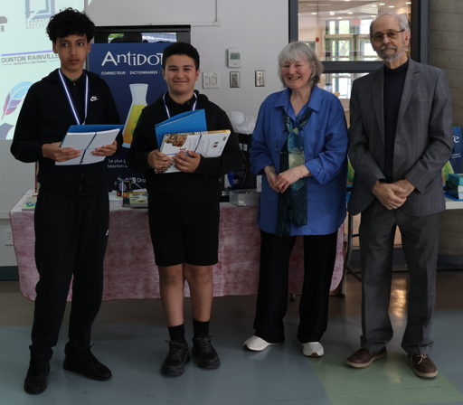 Two students holding certificates and notebooks stand with two adults at a school awards event, smiling by a display table and banner in the background.