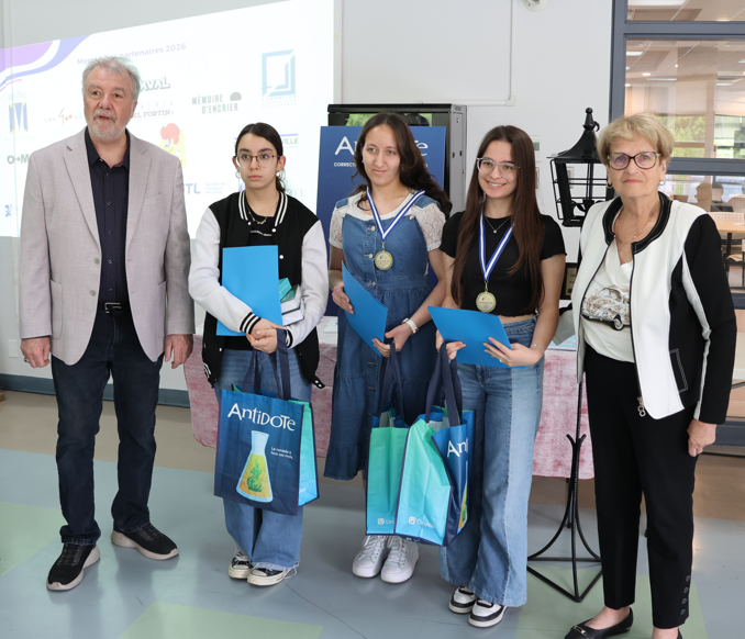 Five people pose indoors for an award ceremony: two adults on either end and three young girls in the center wearing medals and holding blue folders, with gift bags in hand and sponsor logos in the background.