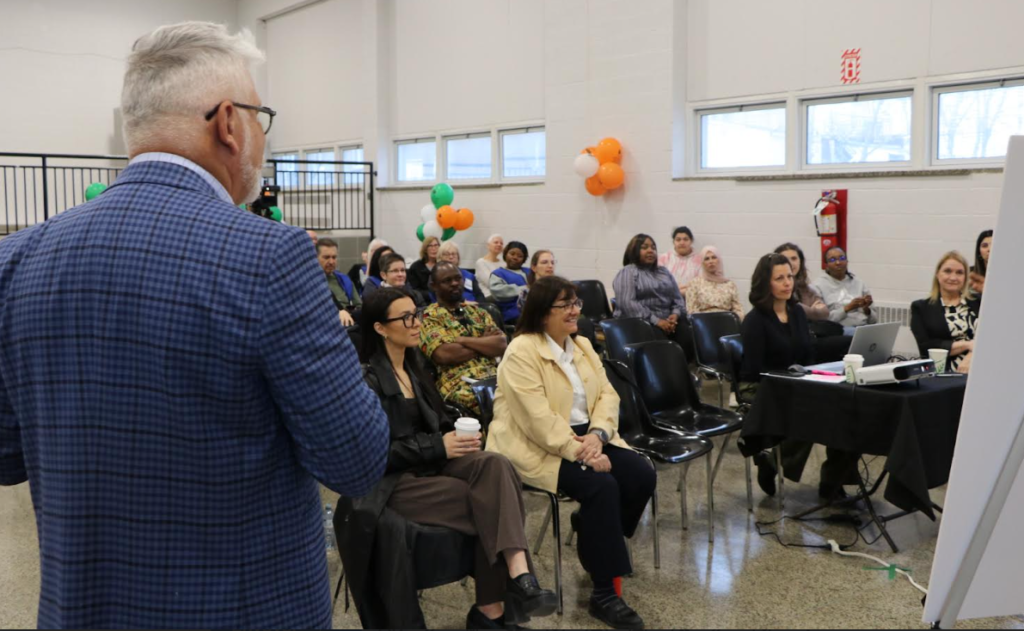 Speaker in a blue checkered blazer addresses a diverse audience in a bright event room with balloons and a projector setup nearby.