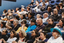 Audience members seated in blue theater seats, watching a presentation at a conference or event.