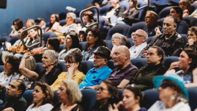 Audience members seated in blue theater seats, watching a presentation at a conference or event.