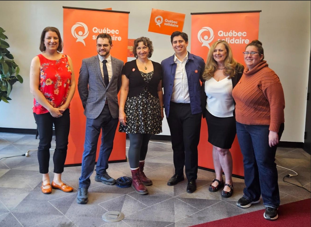 Six adults pose for a group photo indoors with orange Québec solidaire banners in the background.