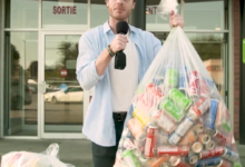 Presenter outside a storefront holding a microphone and a large bag of aluminum cans for recycling.