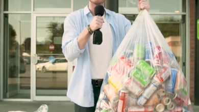 Presenter outside a storefront holding a microphone and a large bag of aluminum cans for recycling.
