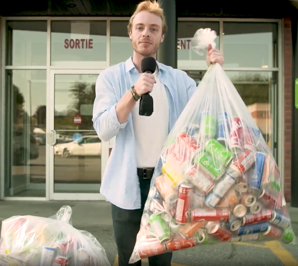 Presenter outside a storefront holding a microphone and a large bag of aluminum cans for recycling.
