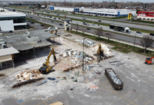 Demolition site in a shopping-center lot with excavators clearing rubble and a debris-filled dumpster nearby, highway in the background