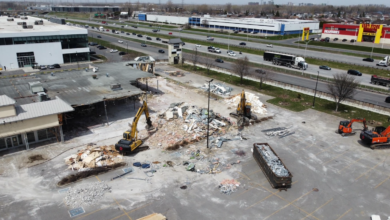 Demolition site in a shopping-center lot with excavators clearing rubble and a debris-filled dumpster nearby, highway in the background