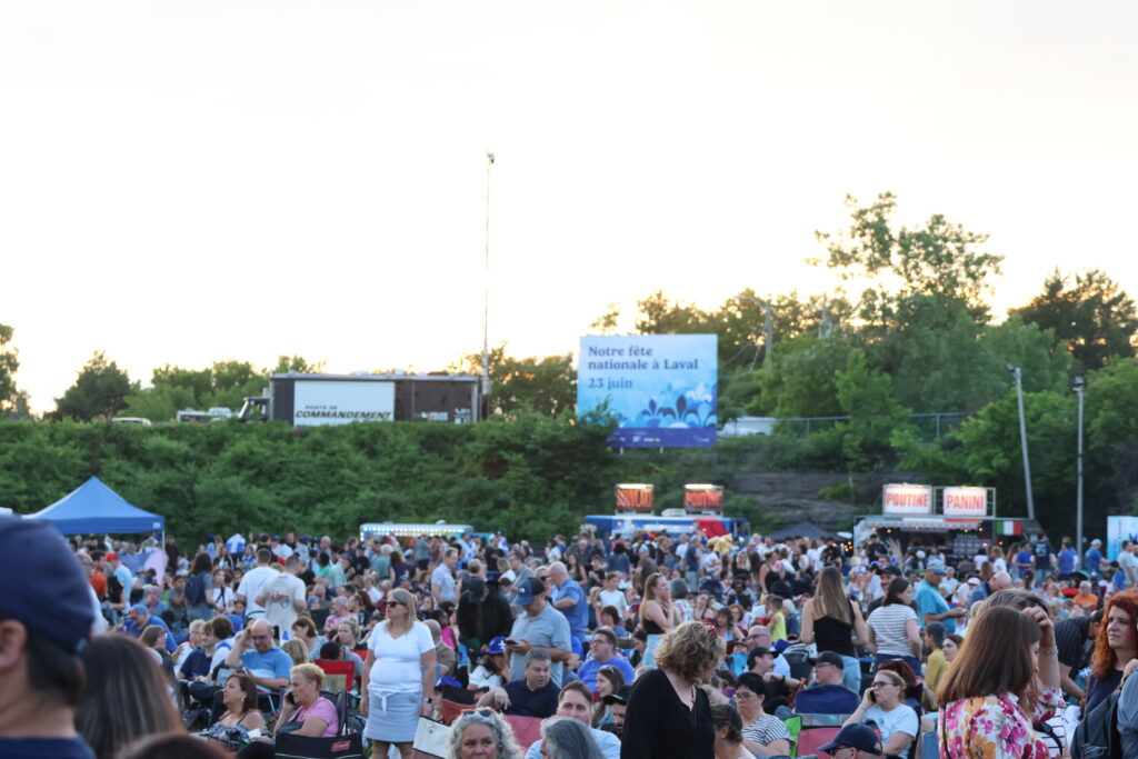 Large crowd gathered at an outdoor festival or event with many people seated and standing, banners and food stalls in the background.