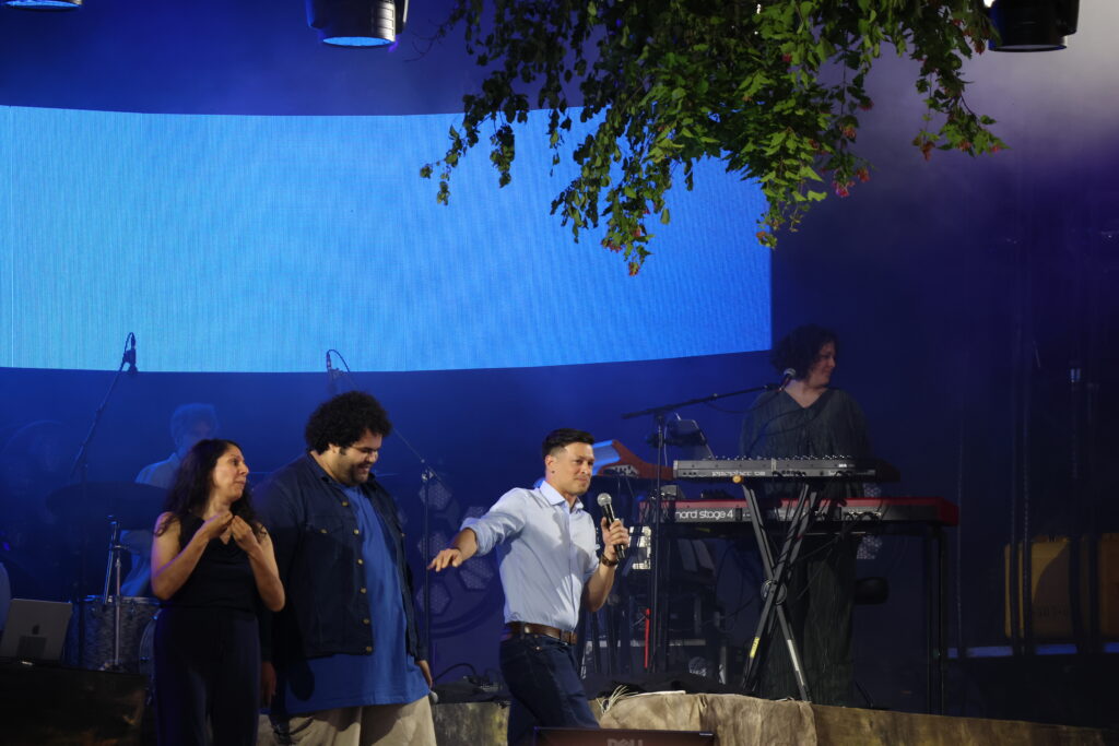 A group of performers on a stage with blue LED backdrop, singing or talking into microphones, with keyboards and other equipment visible behind them.