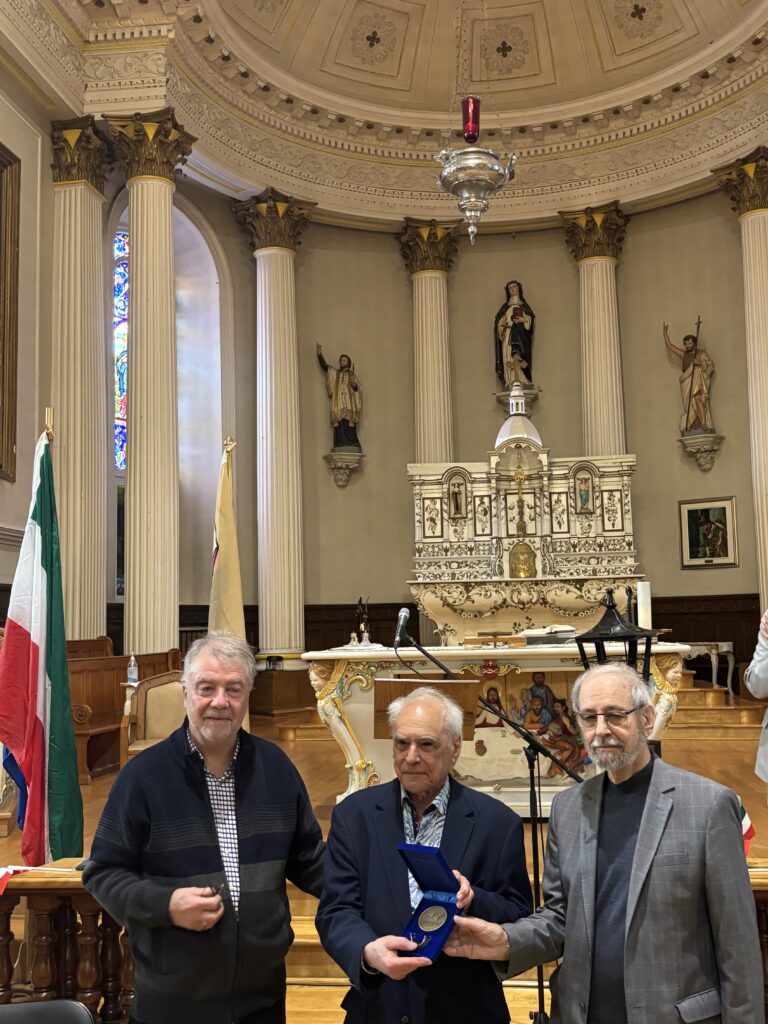 Three men in jackets stand in a church with ornate columns and statues behind them; one holds an open medal case for an award ceremony windowing the altar.