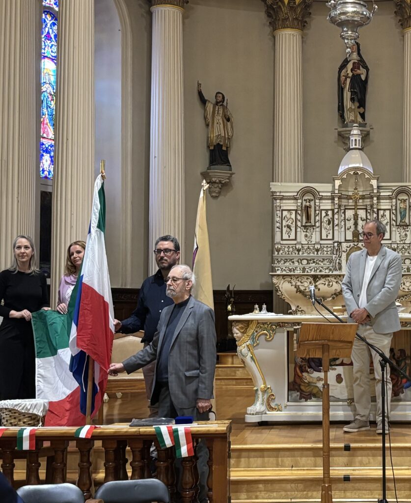 Group of people in a church near an altar, some holding Italian flags with green, white, and red ribbons visible at the front.