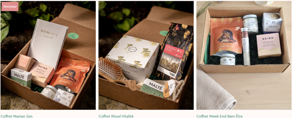 Open gift box with assorted natural skincare and wellness items, including a gray 'MOMENTS' box and pink BKIND packaging on a plant background.