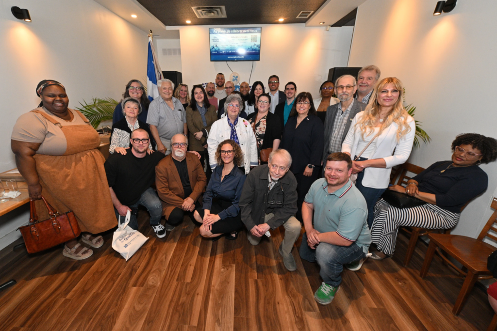 Fête nationale lancement 2026 Média Laval Group of about 25 people posing for a photo in a room with wooden floor and warm lighting, smiling at the camera.