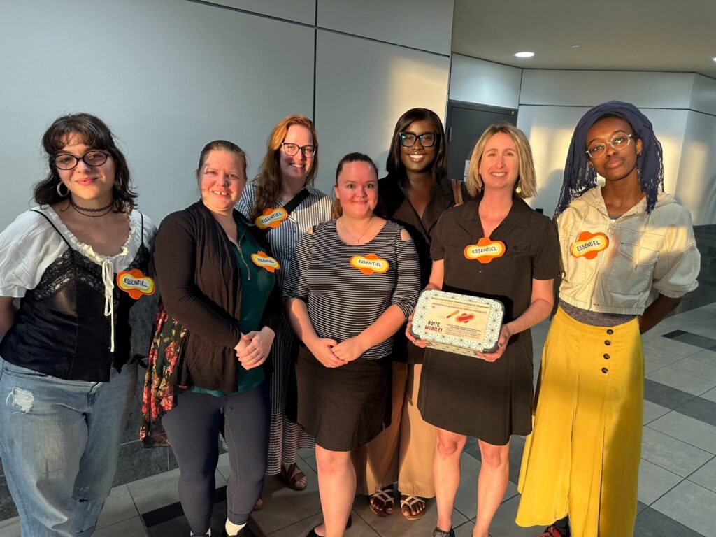 Laval offrira graduellement des produits menstruels gratuits dans certains lieux publics Média Laval Seven women stand together indoors, smiling at the camera, wearing orange ESSENTIEL badges on their chests.