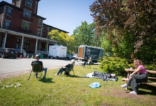 People sit on folding chairs and a bench in a grassy area outside a brick building, with trucks in the background and one person lying under a blanket on the ground near trees.